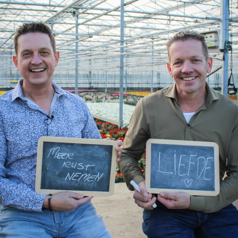 twee mannen in een kas met een krijtbordje in hun handen